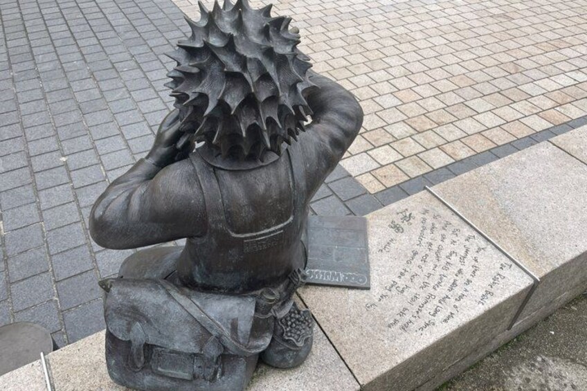 A statue of a person with a spiky head, sitting on a bench with a plaque beside it. 4
