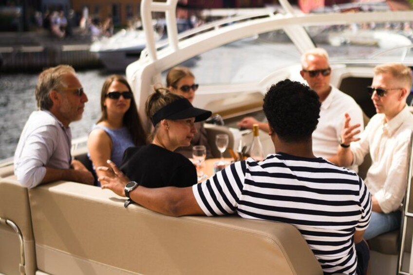 Een groep mensen op een boot, in gesprek, met een jachthaven en andere boten op de achtergrond. 3