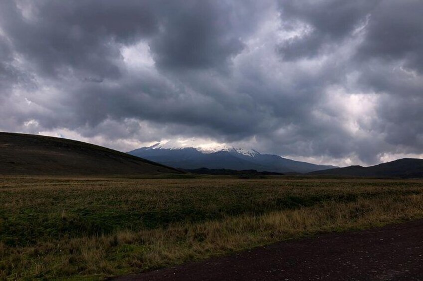 A grassy field with a road, rolling hills, and a mountain range under a cloudy sky. 2