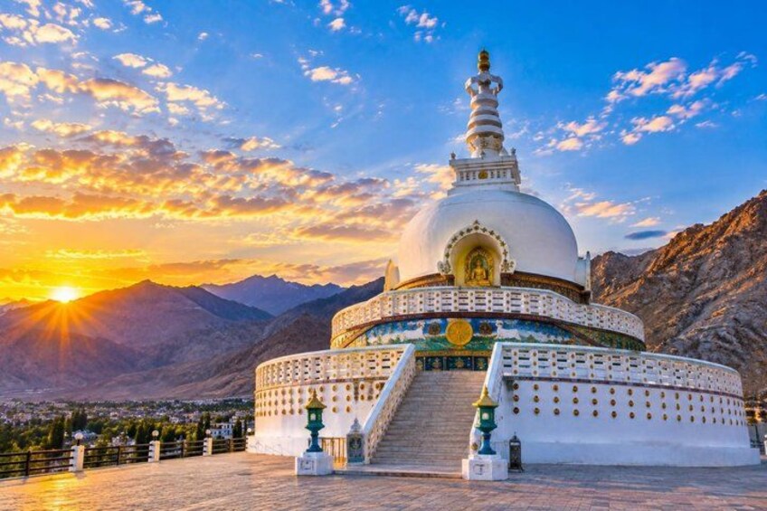A white stupa with a golden spire, surrounded by a white fence, set against a mountainous backdrop with a sunset. 3