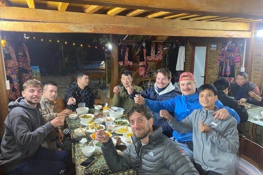 Un grupo de personas sentadas alrededor de una mesa con comida, en un entorno al aire libre con vigas de madera y luces colgantes. 5