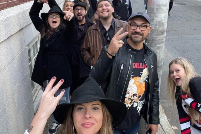 A group of people standing on a sidewalk, some wearing hats and jackets, with a building and a tree in the background. 5