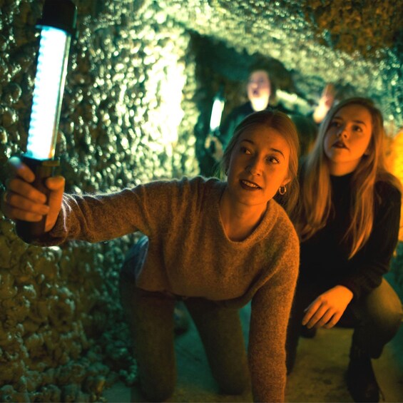 A person holding a smartphone, another person crouching behind, and a stone wall in the background. 1