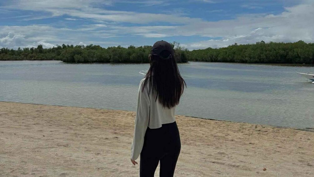 Een persoon staat op een zanderig strand, kijkt uit over een rustig wateroppervlak, met bomen op de achtergrond. 1