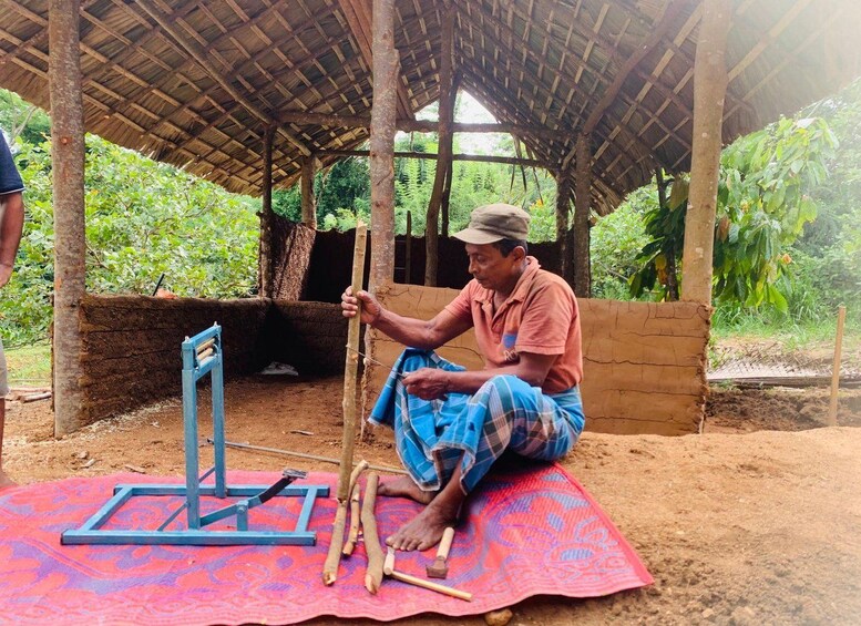 A man sitting on a mat under a thatched roof, working with wooden tools. 1