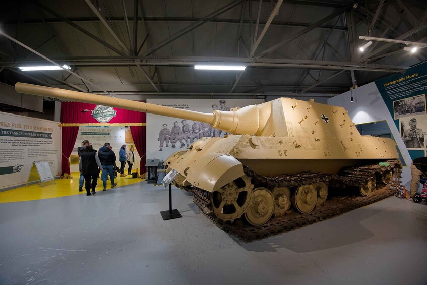 A large tan-colored tank on display in a museum with a red curtain and informational posters in the background. 1