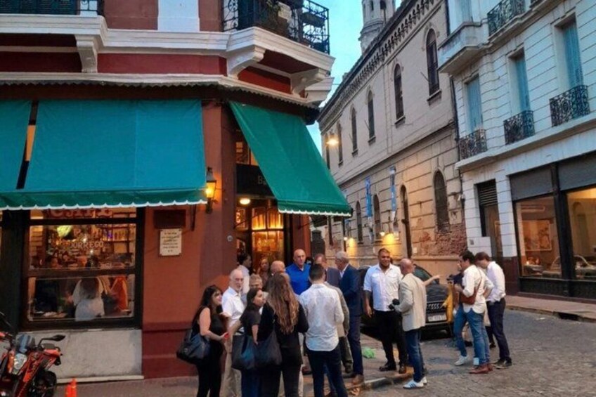 A group of people standing on a cobblestone street in front of a building with a green awning. 4