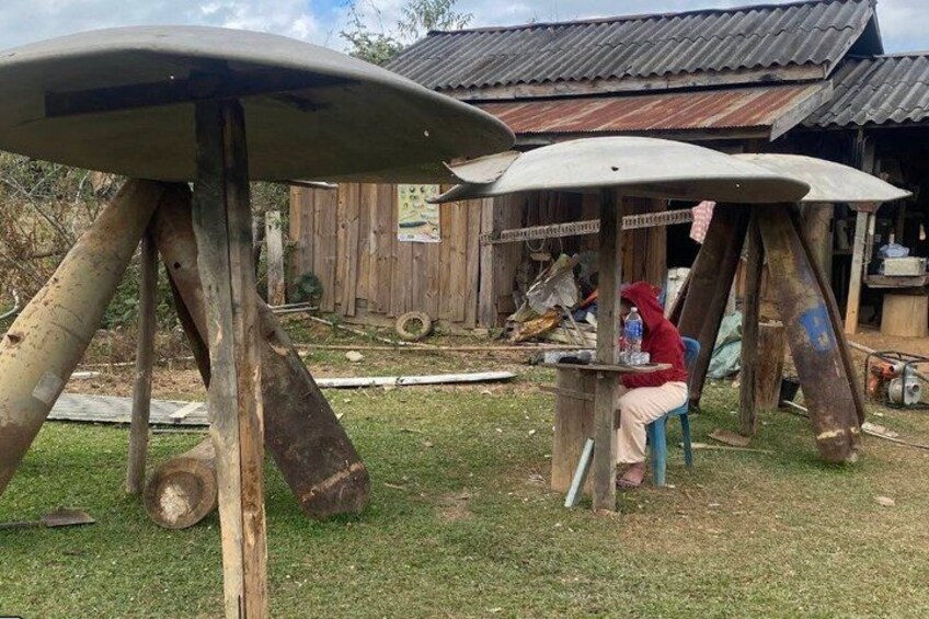 A rustic outdoor setting with a wooden structure, a person sitting under a makeshift shelter, and a surfboard on a stand. 5