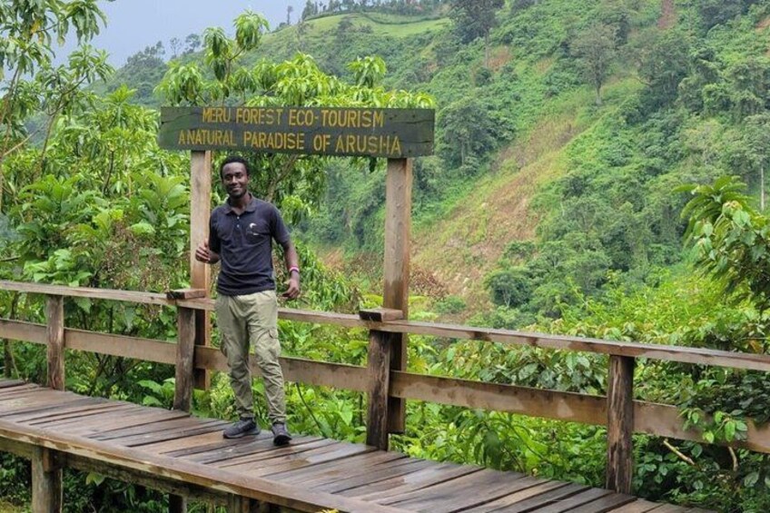 A man standing on a wooden bridge with a sign that reads 'Meru Forest Eco-Tourism Anamath of Arusha.' 3