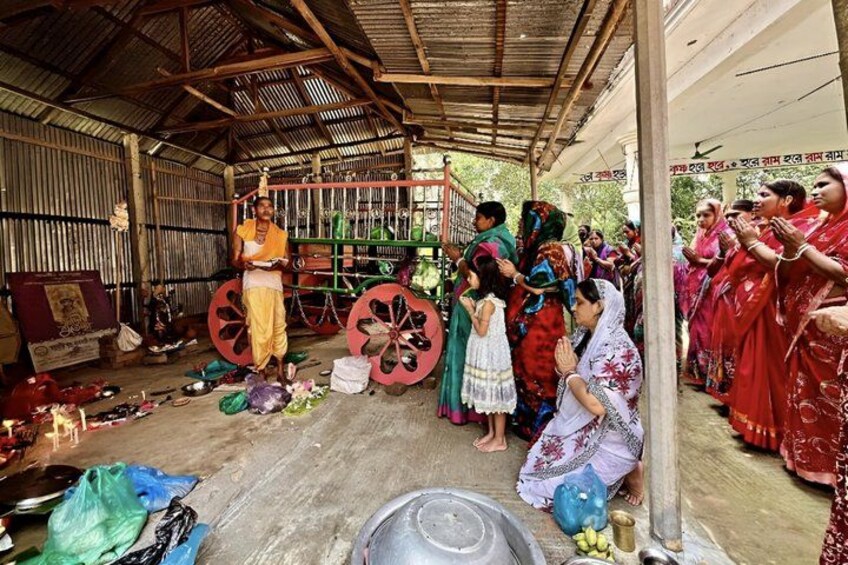 A group of people in traditional attire gathered in a rustic indoor setting with wooden structures and a makeshift stage. 4