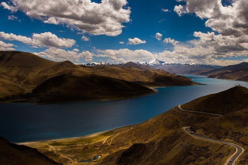 A mountainous landscape with a winding road, a large lake, and distant snow-capped peaks. 5