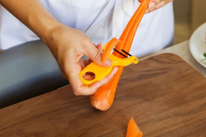 A person using a manual peeler to peel a carrot on a wooden cutting board. 3