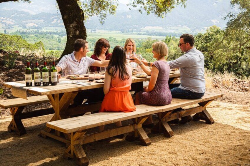 A group of people dining at a wooden picnic table outdoors with a scenic view in the background. 2