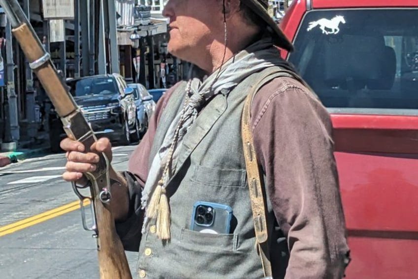A person holding a rifle, wearing a vest with a phone in a pocket, standing on a street with cars and buildings in the background. 5