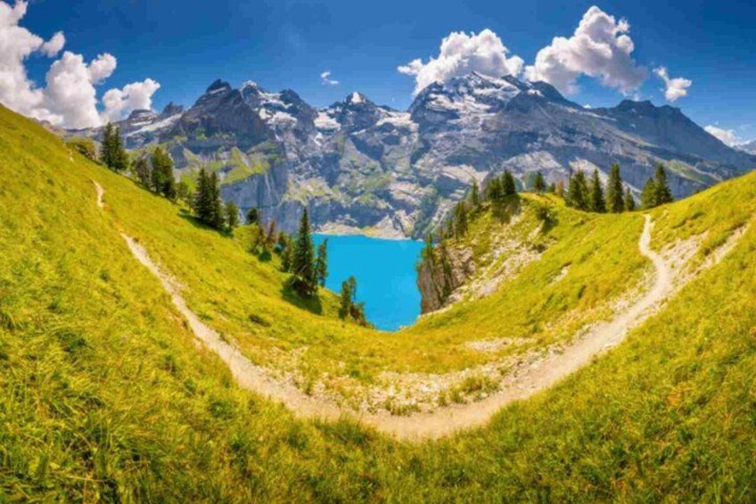 A mountain landscape with a clear blue lake, a hiking trail, and snow-capped peaks. 4