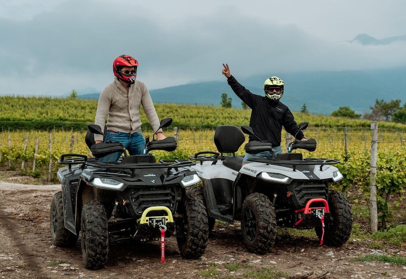 Two people on ATVs in a vineyard with mountains in the background. 1
