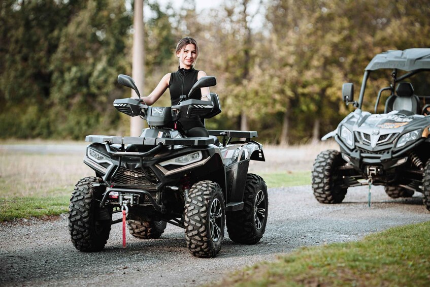 A person sitting on a black ATV with a red tow rope, parked on a gravel path with another ATV in the background. 3