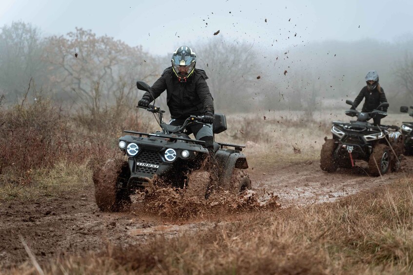 Two riders on ATVs on a muddy trail with dry grass on either side. 5