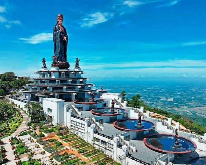 A large statue on a multi-tiered platform with fountains, surrounded by a landscaped garden and overlooking a coastal view. 4