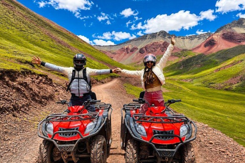 Two people on ATVs on a dirt road with green hills in the background. 1