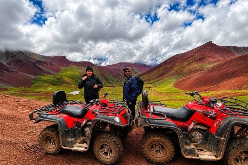 Two people standing next to red ATVs on a dirt path with mountains in the background. 2