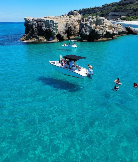 A clear blue sea with a rocky coastline, a boat with people on it, and swimmers in the water. 5