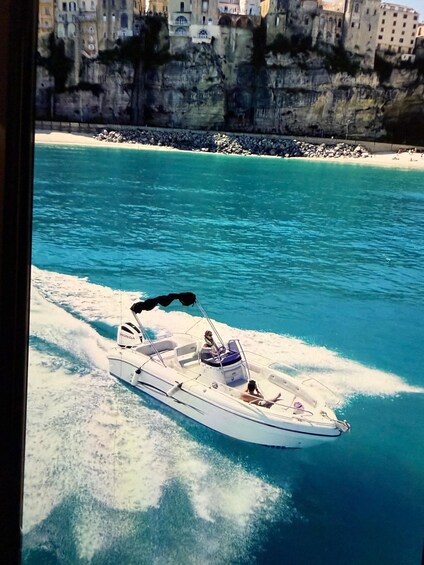 A boat on a turquoise sea with a rocky coastline and buildings in the background. 3