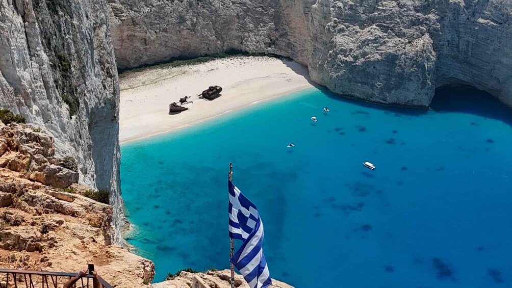 A coastal landscape with a clear blue sea, rocky cliffs, a sandy beach, and a Greek flag. 5
