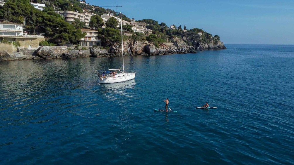 A sailboat on calm waters, a person paddleboarding, and a coastal town with buildings on a hillside. 4