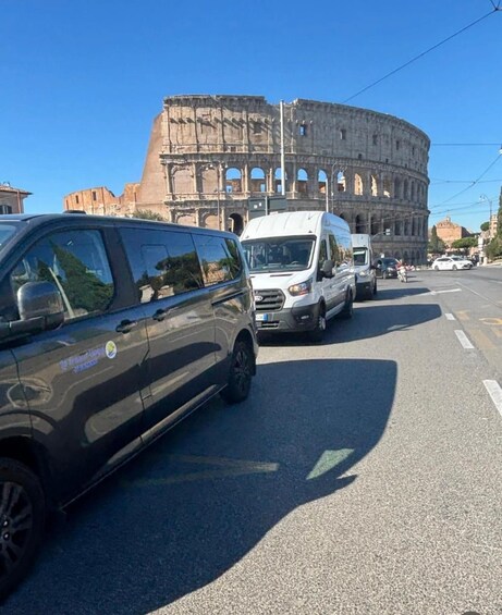A busy street with vehicles, including a black car and a white van, in front of the Colosseum. 5