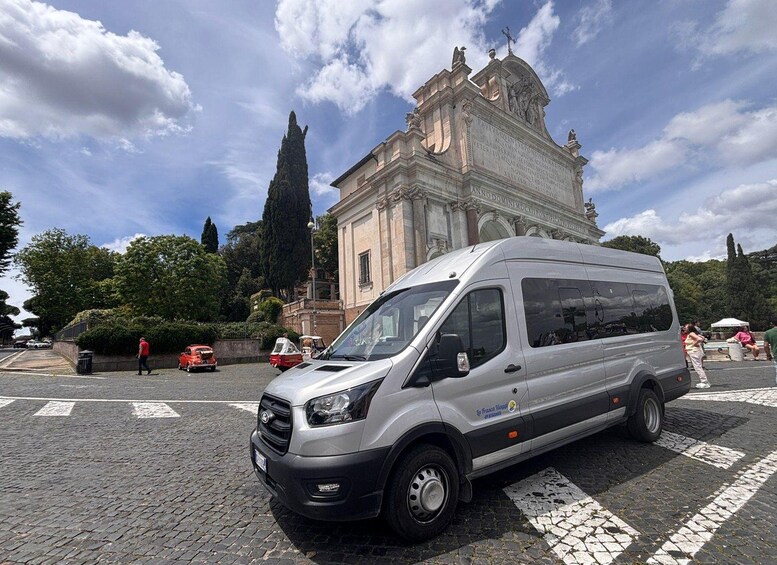 A silver van parked on a cobblestone street with a historic building in the background. 1