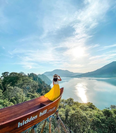Eine Frau im gelben Kleid steht auf einem hölzernen Boot mit der Aufschrift 'hidden hill cruise' und blickt auf einen ruhigen See mit Bergen im Hintergrund. 5