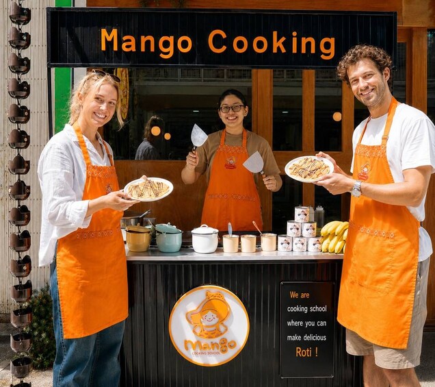 A cooking school named Mango Cooking with two instructors holding plates of food, a signboard, cooking utensils, and ingredients. 1