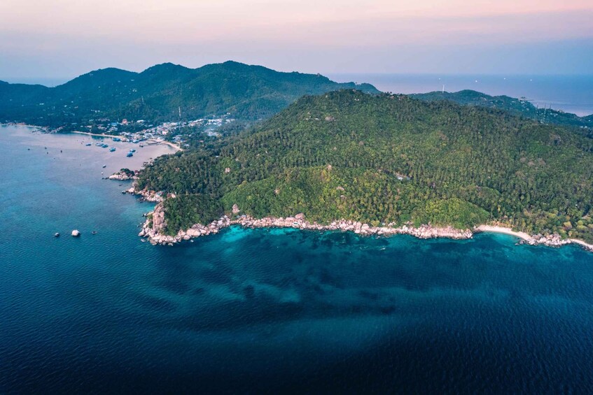 Vista aérea de una zona costera con una pequeña isla rodeada de aguas azules cristalinas, una playa arenosa y una extensa cadena montañosa cubierta de bosques al fondo. 5