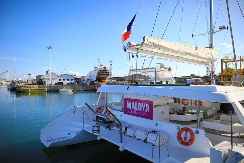 Una barca bianca di nome MALOYA LA REUNION ormeggiata in una marina, con un cielo blu limpido e edifici industriali sullo sfondo. 2