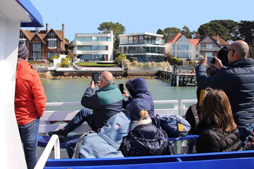 People on a boat, some taking photos, with modern houses and a river in the background. 5