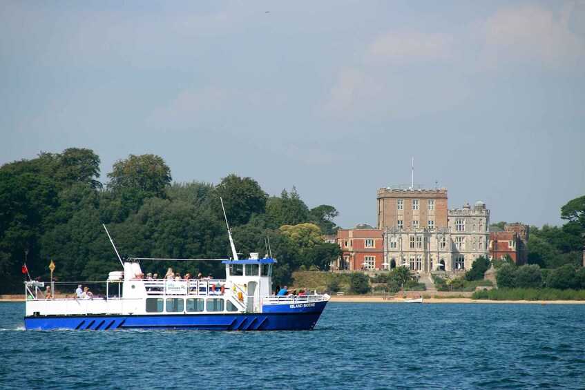 A blue and white boat with passengers on a calm body of water, a large historic building in the background, and a forested shoreline. 4
