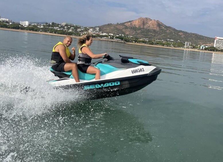 En person kører jetski på roligt vand med et bjerg og bygninger i baggrunden. 4