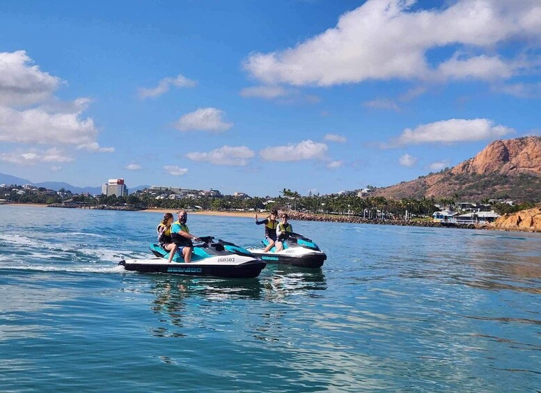 To jetski'er på roligt vand med klar himmel, en klippefyldt kystlinje og en bygning i det fjerne. 2