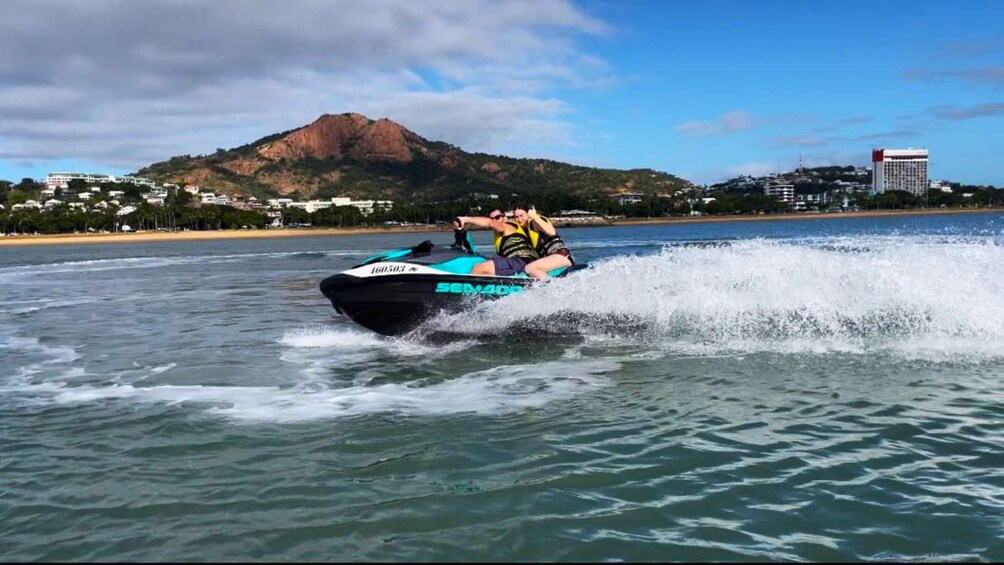 En person på jetski på vandet, med et bjergrigt landskab og bygninger i baggrunden. 5