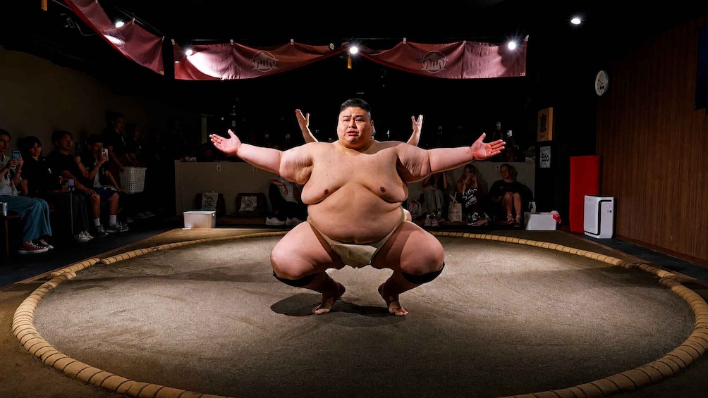 A sumo wrestler in a squat position on a stage with an audience in the background. 2
