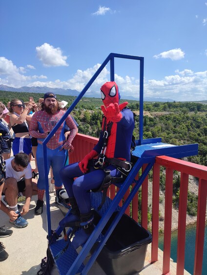 A person in a Spider-Man costume sitting on a blue and red metal structure with a scenic view of mountains and greenery in the background. 5