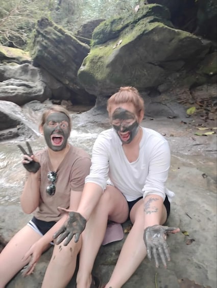 Two people sitting on a rocky shore with moss-covered rocks and a river in the background. 4
