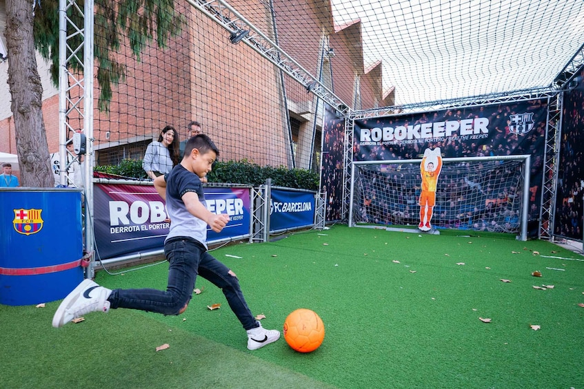 A person kicking a soccer ball on artificial turf with a goal and a banner in the background. 2