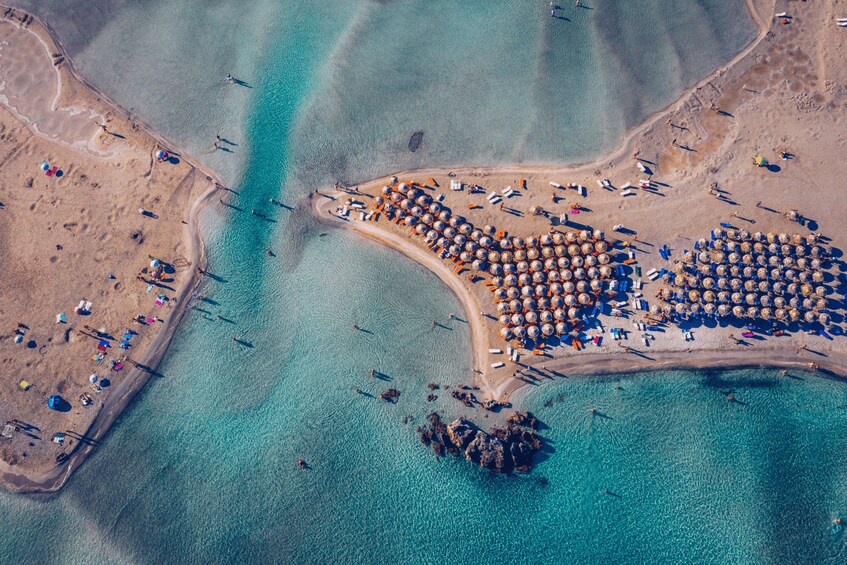 Eine Vogelperspektive zeigt einen Strand mit klarem türkisblauem Wasser, einem sandigen Ufer und zahlreichen Sonnenschirmen und Liegestühlen. 4