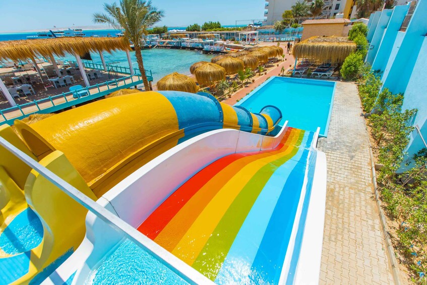 A water slide with a rainbow design, a pool, and a beach area with umbrellas and lounge chairs. 5