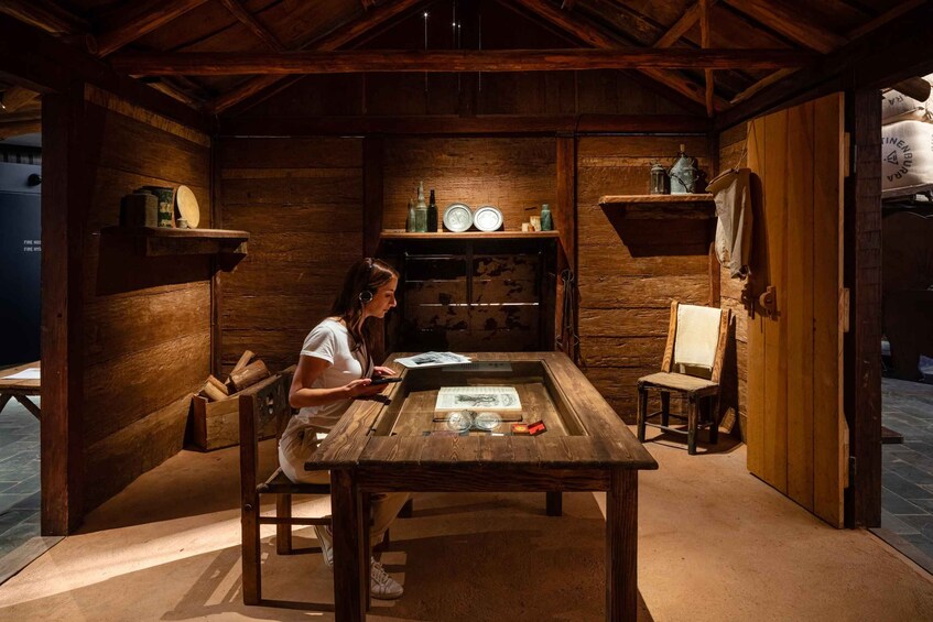 Ein rustikales Holzinterieur mit einer Frau, die an einem Tisch sitzt und ein Buch liest. An der Wand befinden sich Regale mit Flaschen und ein Laternen. 2