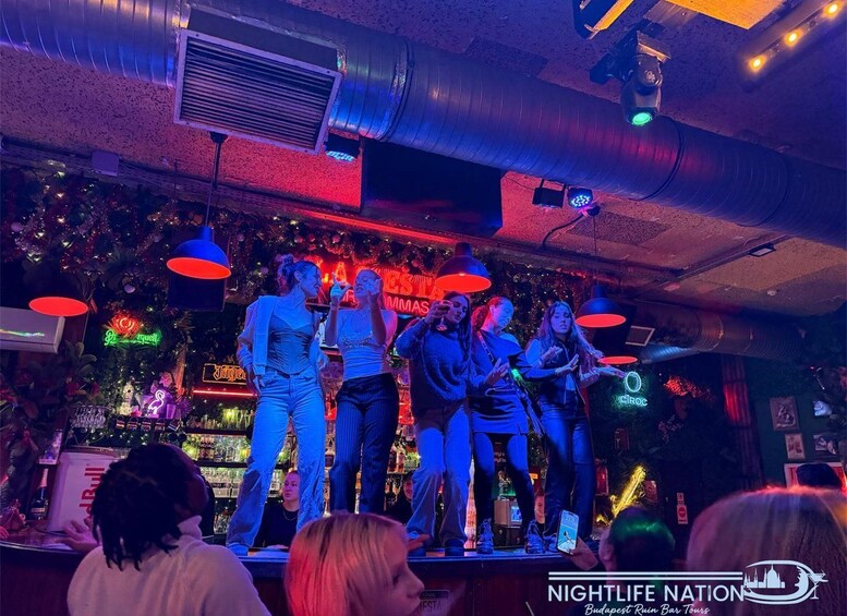 A bar with a stage where people are performing. There are neon signs and a bar counter visible. 4