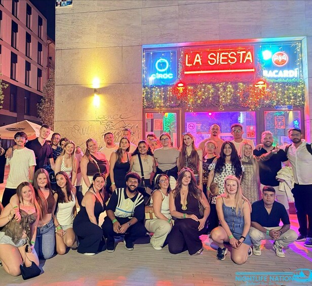 A group of people posing in front of a building with illuminated signs reading 'LA SIESTA' and 'BACARDI'. 1