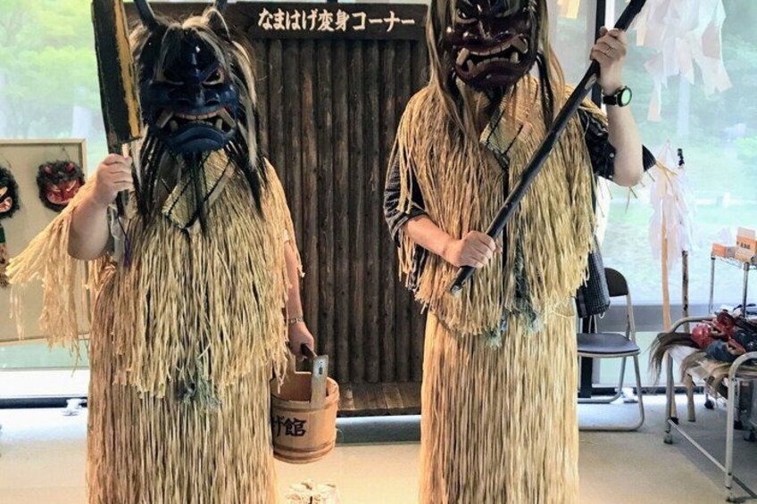 Two individuals in traditional straw costumes holding staffs, standing in front of a wooden backdrop with Japanese text. 2